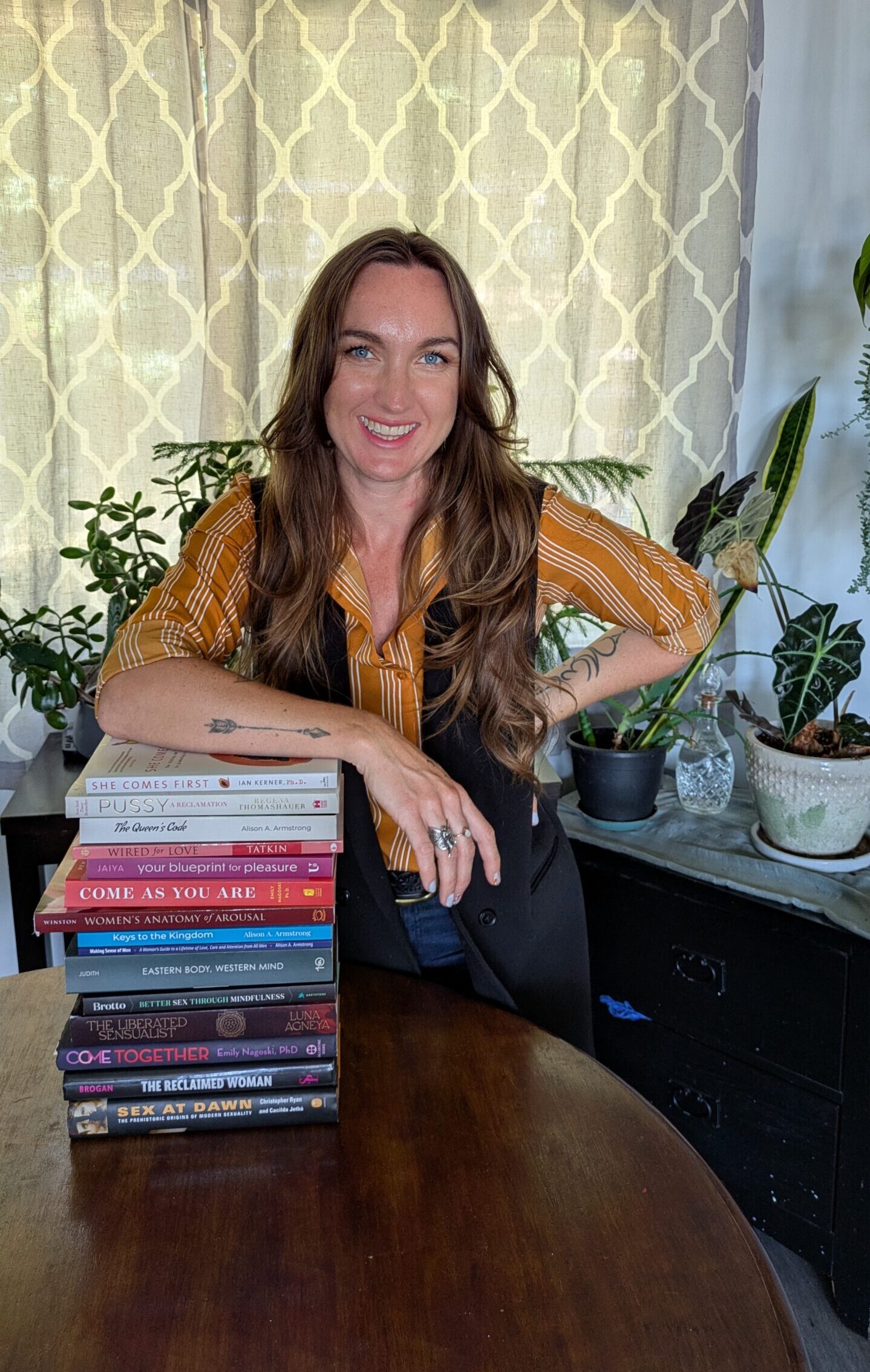 Jannine MacKinnon, certified sex, love, and relationship coach, smiling beside a stack of books on women's pleasure and sexuality