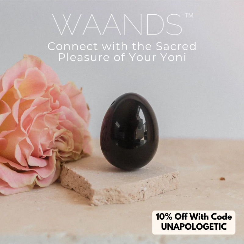 An obsidian yoni egg sits on top of a tan coloured stone slab with a pink rose to the left of it with a coupon code displayed for 10% off Waands Crystal Pleasure Tools with code UNAPOLOGETIC